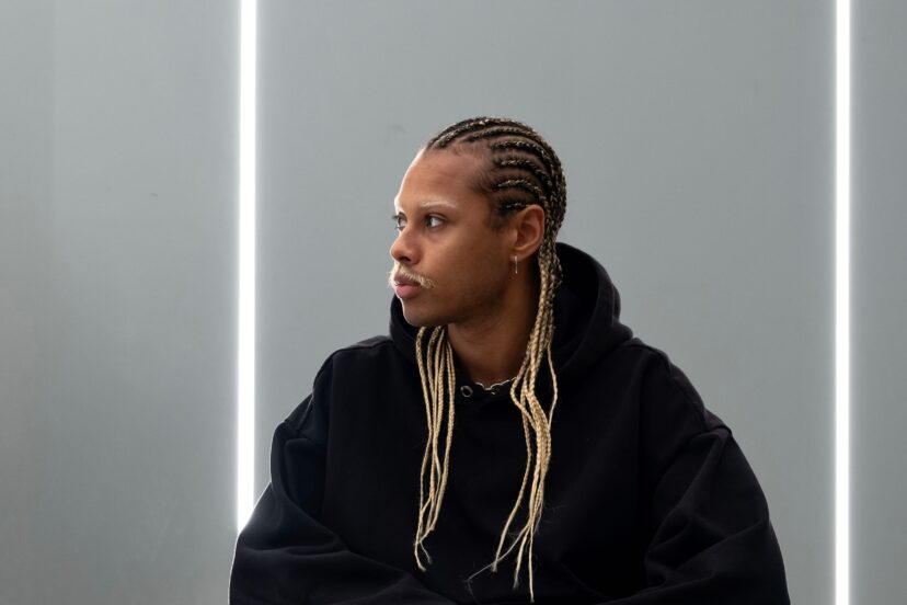 A man looks towards his right. He has a moustache and long braids with blonde in them. He is wearing a big black hoodie. Behind him is a blank wall with a white strip light to his left.