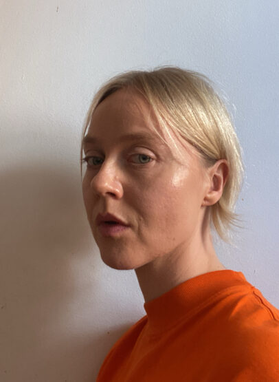 A person with short blonde hair looks at the camera with an angled glance. They are wearing a bright range top and are positioned against a white wall. 
