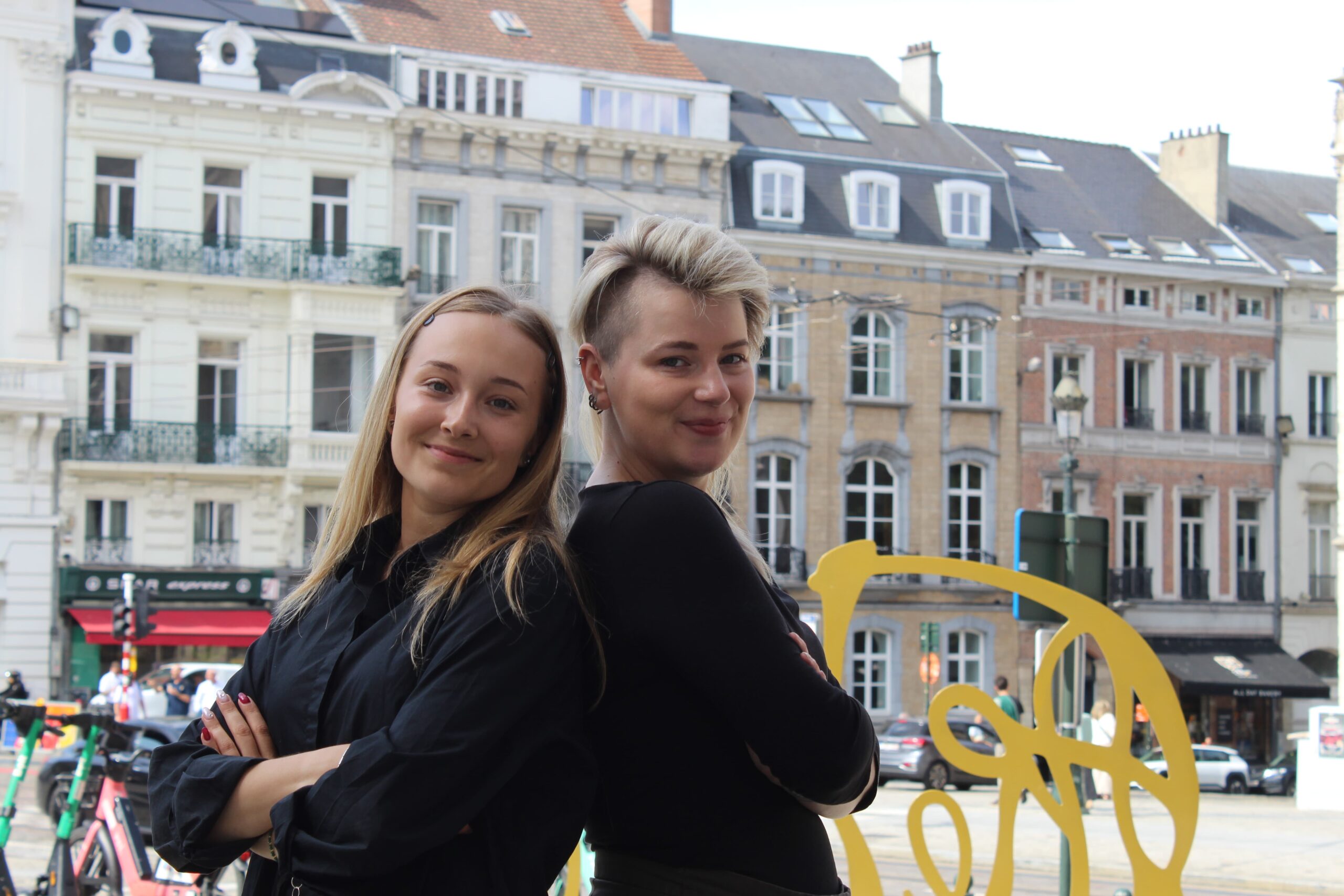 Two people stand back to back and smile at the camera. They are stood in a street, in front of a line of buildings. They are both wearing black.