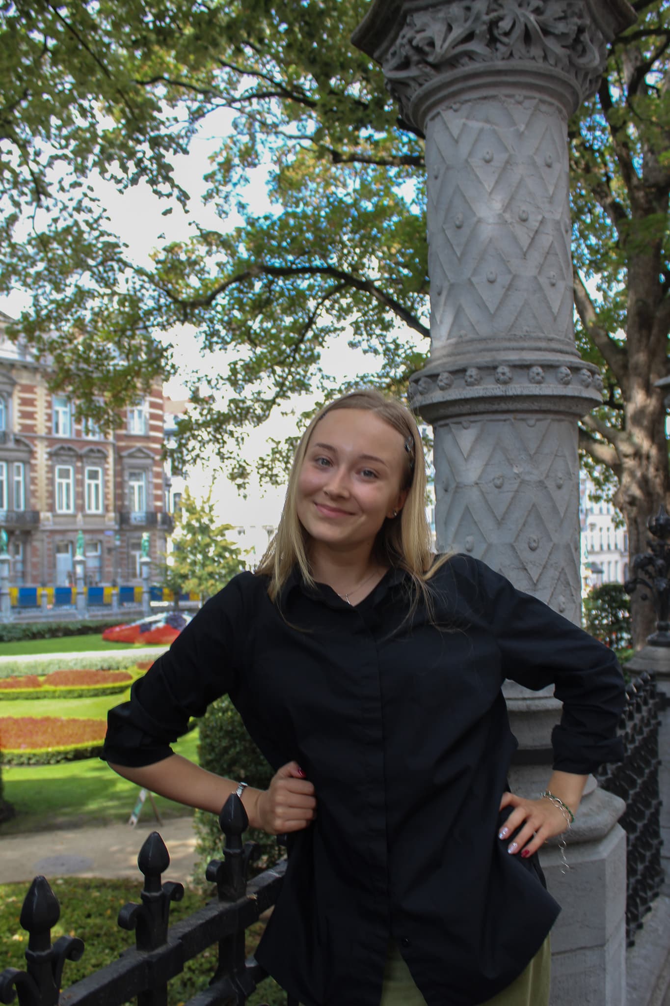 A smiling young woman in black shirt poses beside an ornate stone column and iron fence in a leafy city park, with colorful gardens behind her.