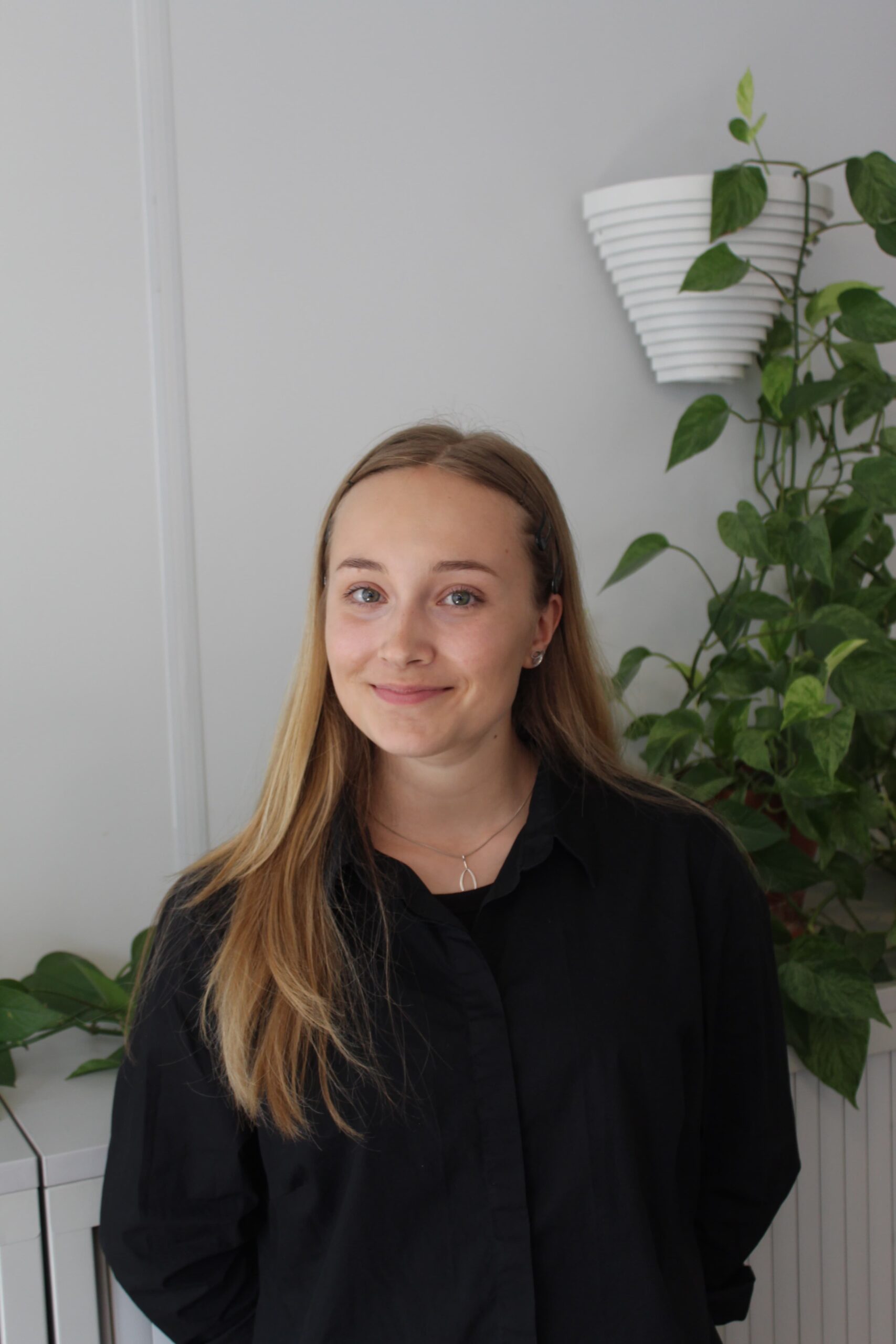 A woman in a black shirt and long light brown hair smiles at the camera. She is standing against a white wall with plants growing down it.