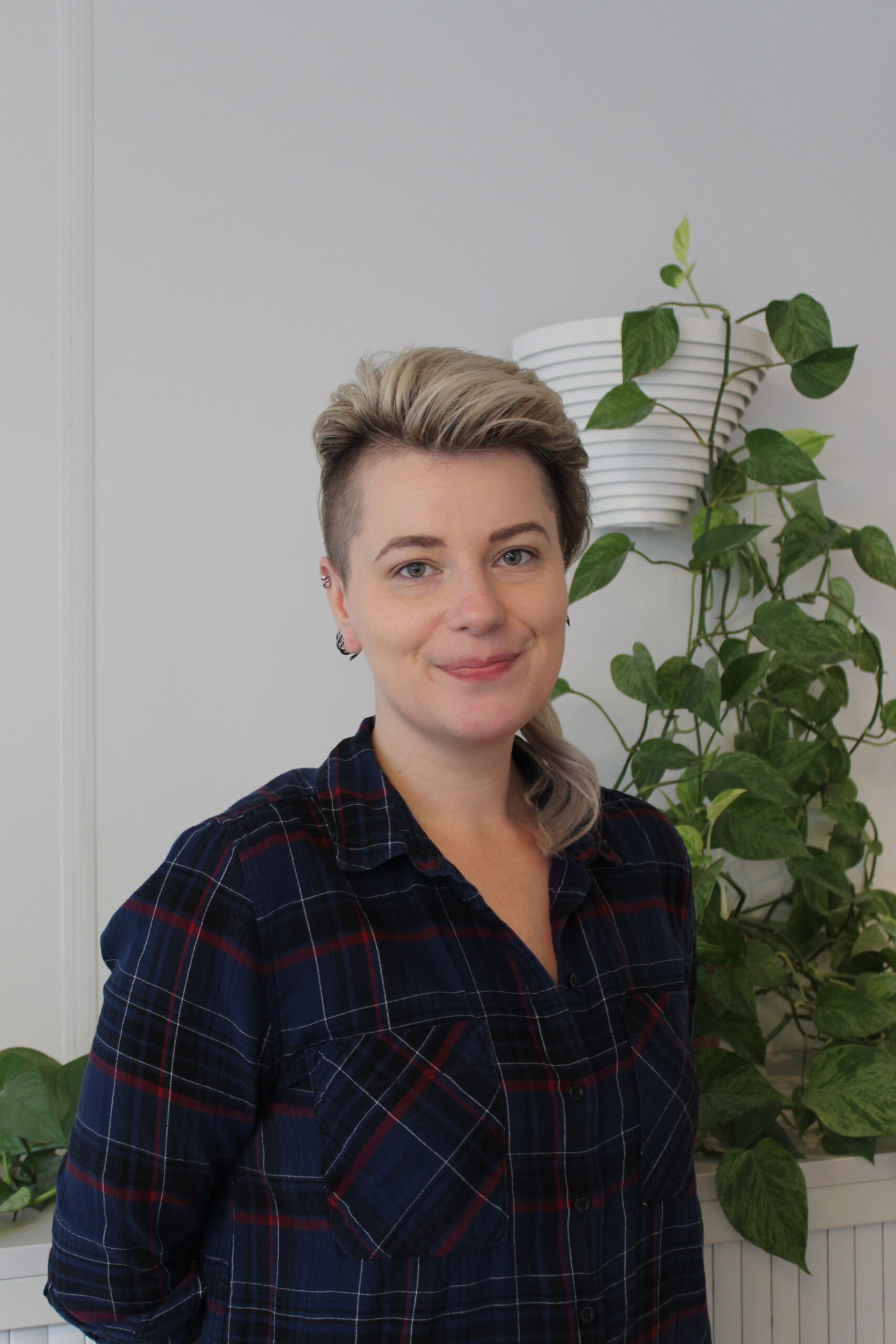 A person in a dark, tartan shirt, with their hair brushed back smiles at the camera.  They are standing against a white wall with a green plant growing down it.