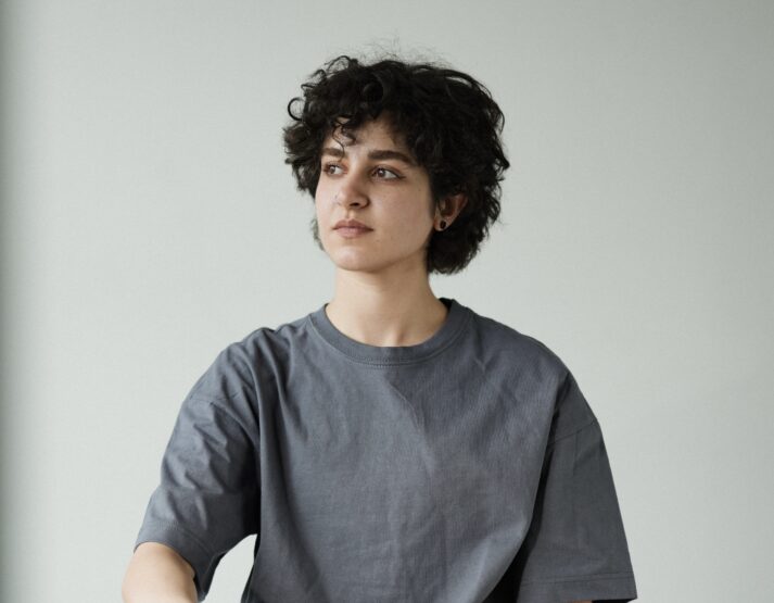 A woman with dark curly hair, a nose ring and earring looks off into the distance. She is waring a baggy grey T-shirt against a blank background.
