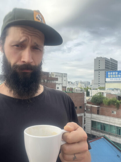 A man with a cap, dark beard and moustache and a black T-shirt looks at the camera. He is holding a white mug, with the view of a city visible behind him.