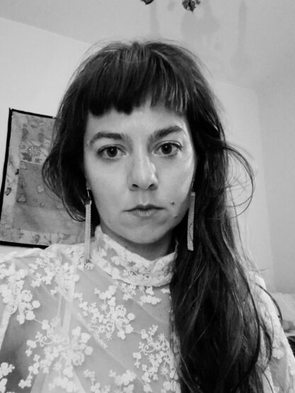 A woman with long her and a fringe looks directly at the camera. The headshot is in black and white. She is wearing a white lace dress with flower details and long earrings.