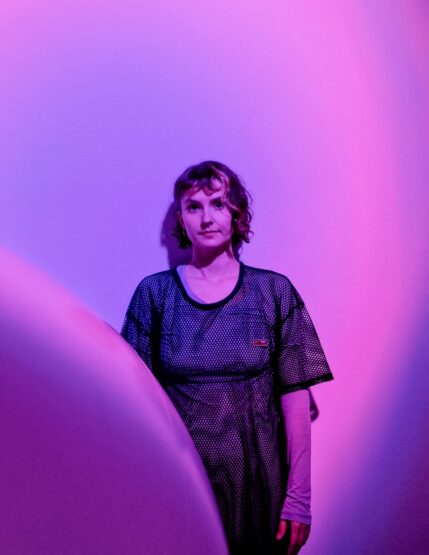 A woman with chin-length hair stands against a wall. The way and the room around her is lit up with purple light, she is also bathed in purple. a large round object partially blocks the view of her.