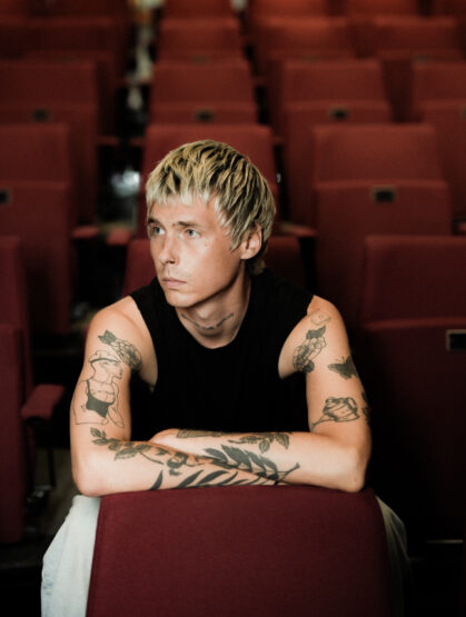 A man sits on a chair with his his arms over the back of the seat. He has a bkacl tank top on, with tattoos on his arms and short dyed blonde hair. Behind rows of red theatre seats can be seen.
