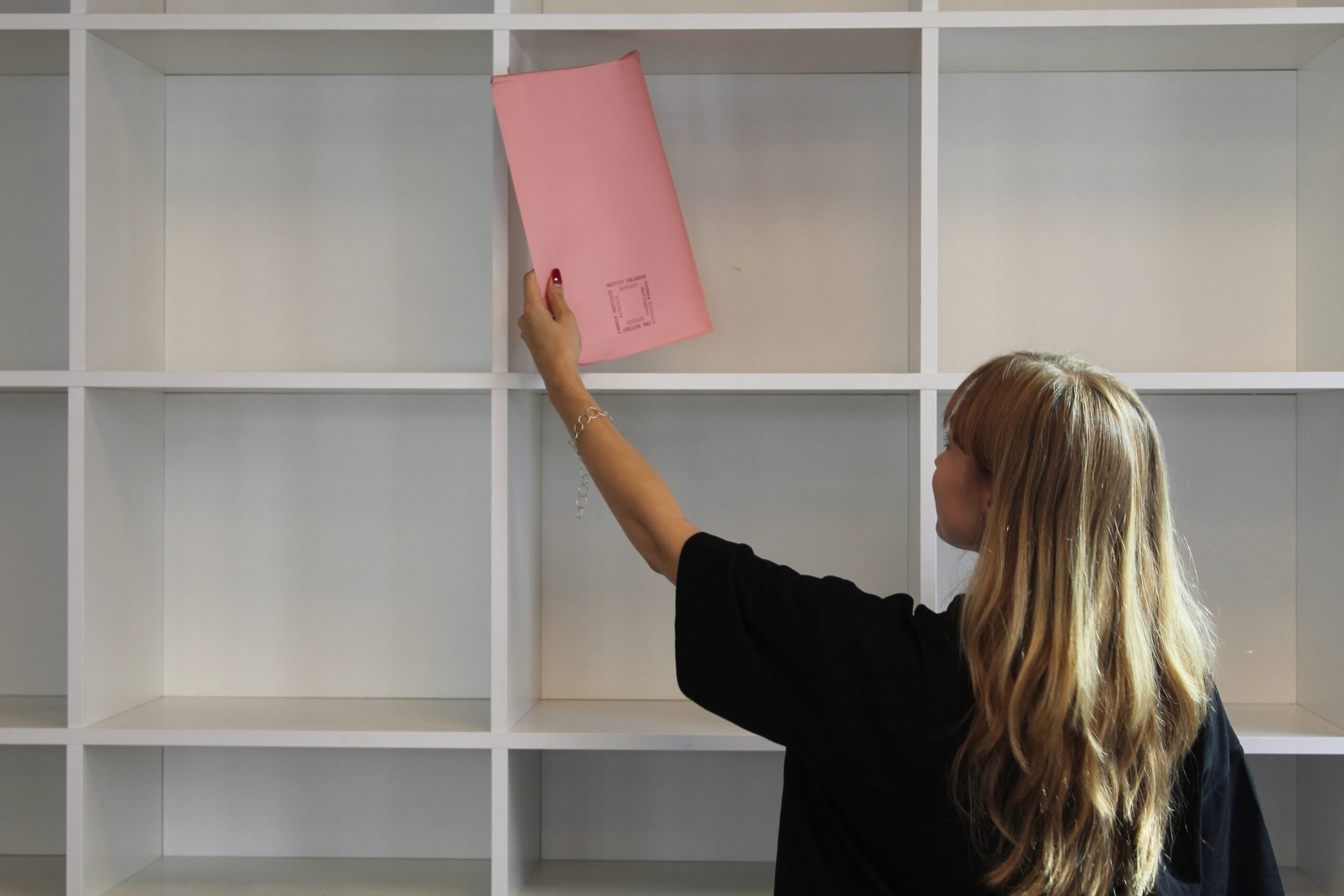 A woman with long brunette hair, wearing a black T-shirt reaches up to take a pink folder from a white shelving unit.