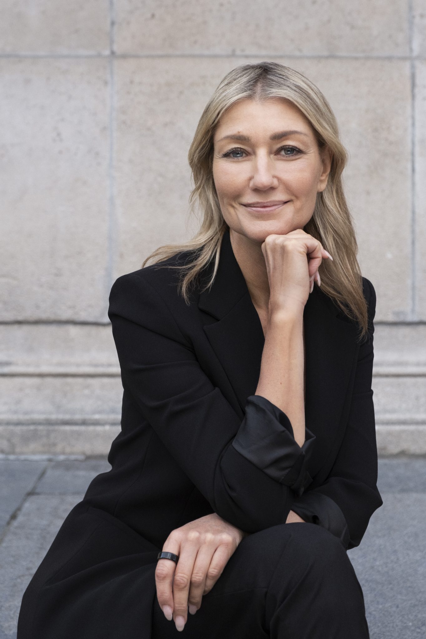 A woman with long blonde hair, wearing a black blazer sits outside a light, stone building. She smiles as she rests her chin on her hand.
