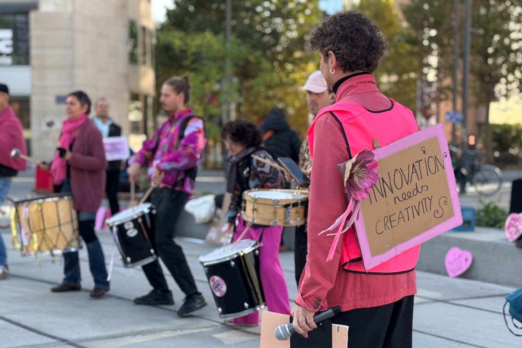 A person dressed in pink with short curly hair standing back towards camera with a group of drummers with a sign saying innovation needs creativity.