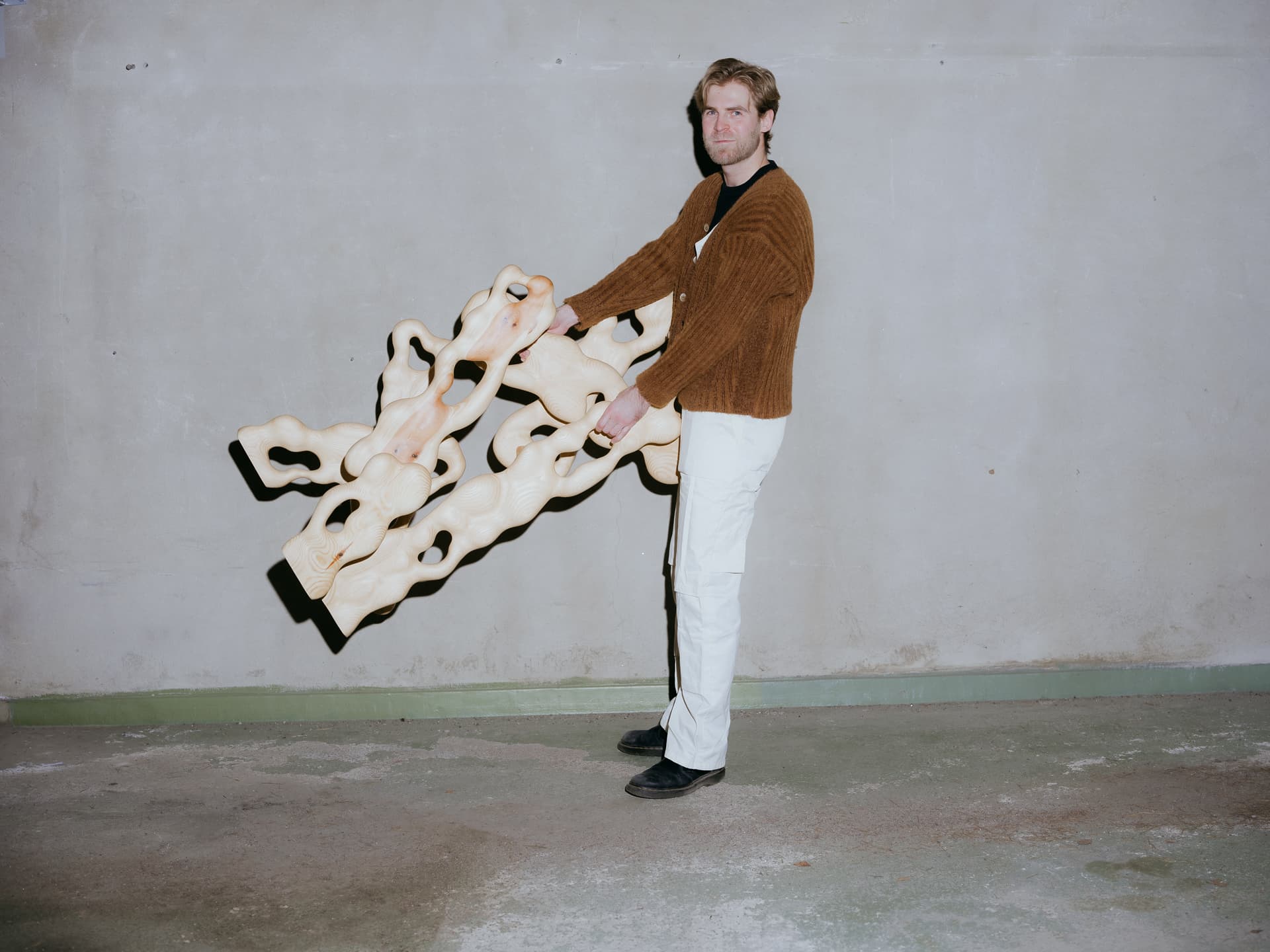 A man stands facing to the left holding sculptures made of pine wood out in front of him. He is wearing a brown jumper and white trousers.