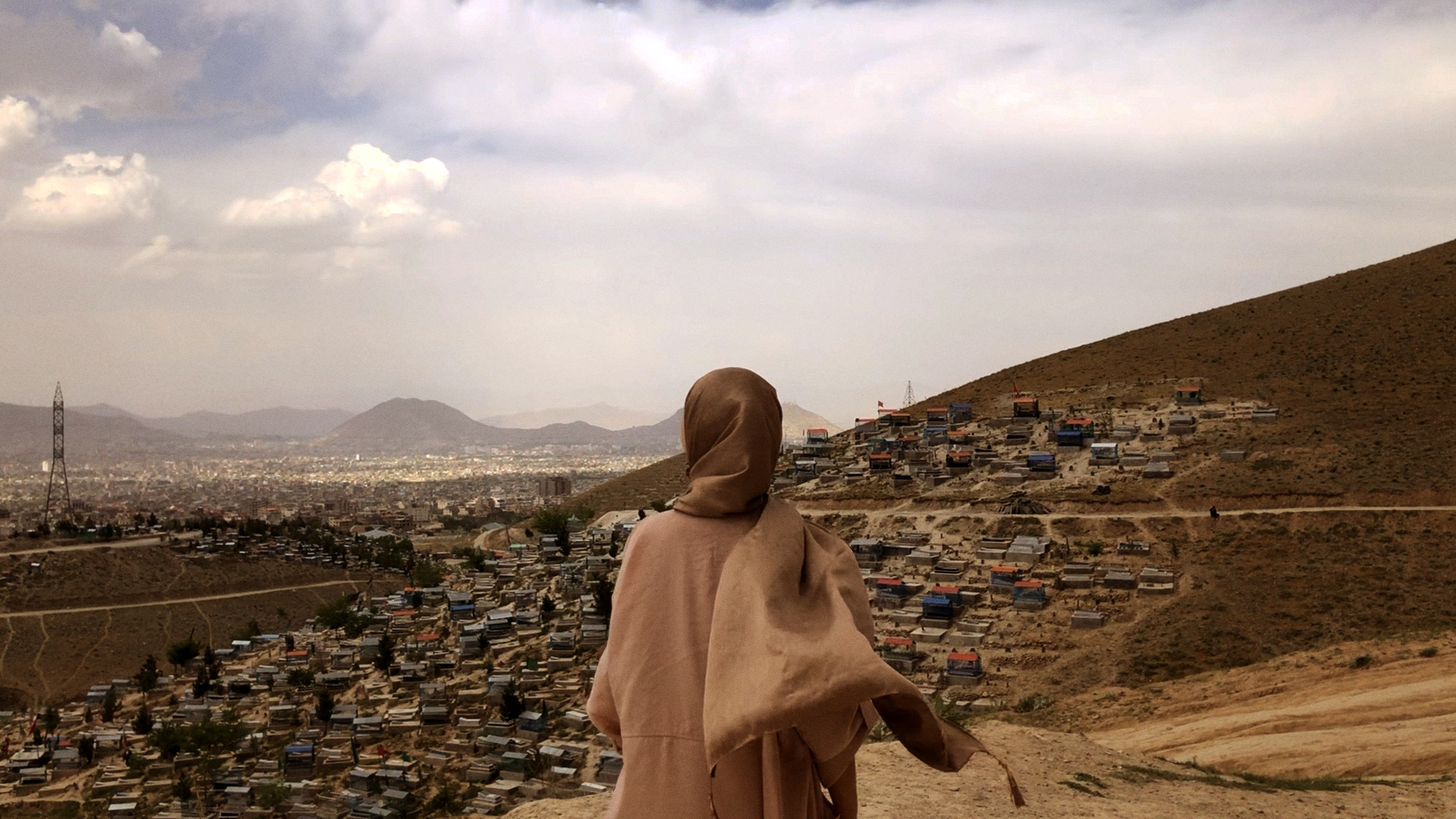 Yellow FIlm & TV The Secret Reading Club of Kabul Still 2 Protagonist Dua in Kabul A person wearing a head scarf looks out to the horizon, over Kabul. The houses are seated in a valley surrounding by hills.